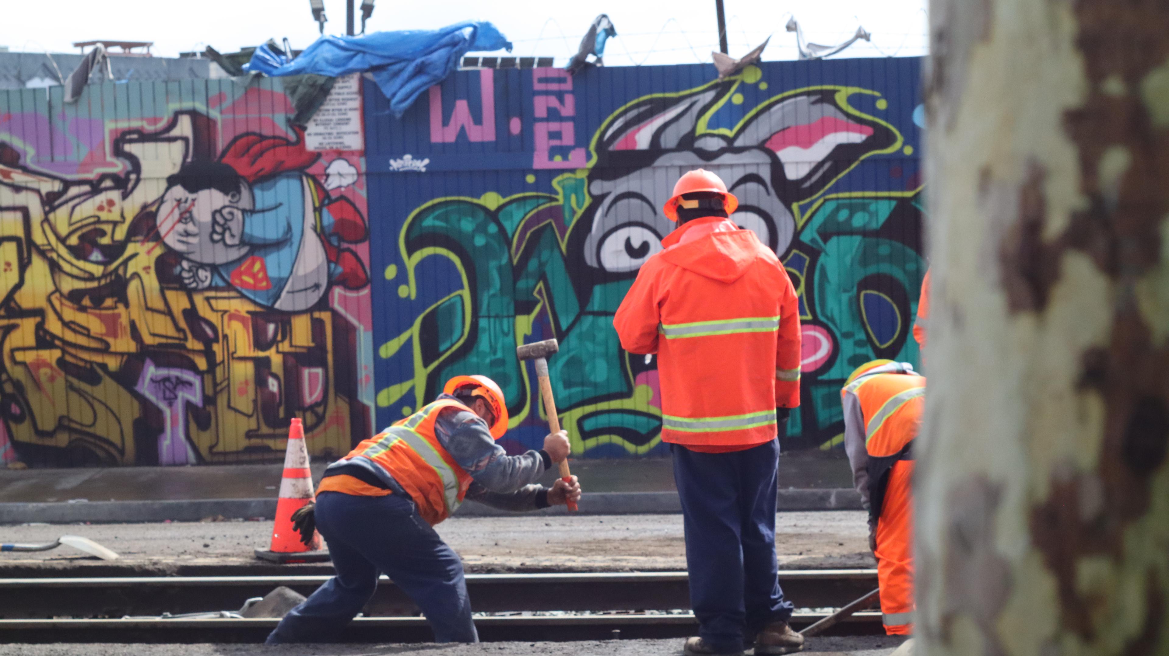 Construction workers along the Orange Line alignment, in front of a wall of graffiti